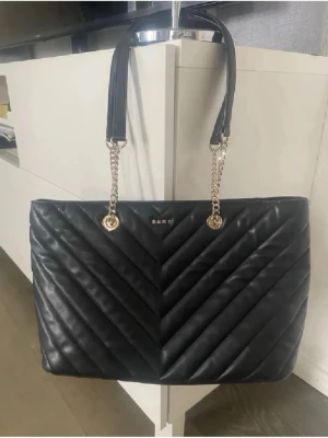 Svart quiltad handväska från DKNY - Snygg svart quiltad handväska från DKNY med guldfärgade kedjedetaljer och axelremmar i läder. Väskan har ett elegant V-format mönster och metallogo framtill. Perfekt för dig som vill ha en stilren och trendig accessoar. Kan va lite sliten vid handtagen men inget som syns eller stör💕
