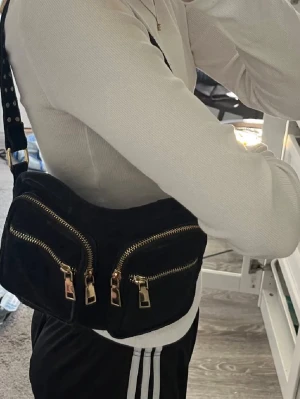 Svart axelväska med nitar och guld-detaljer noella - Snygg svart axelväska i mockaimitation med två stora framfickor och guldfärgade dragkedjor. I fint skick! Säljer då jag har andra väskor och använder ej, inte använd mycket och helt fläckfri inuti