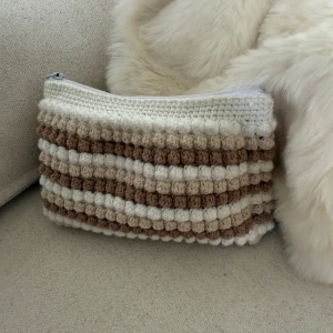 Necessär  - Säljer en handgjord virkad necessär i med beige och vita ränder. Väskan har ett bubbligt mönster och stängs med dragkedja upptill. Perfekt accessoarer och små grejer.