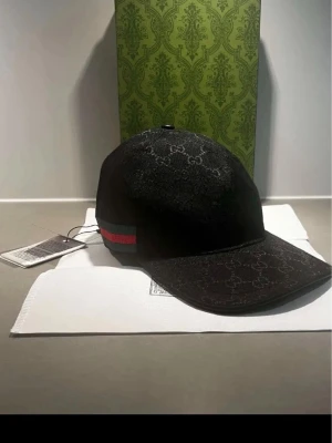 Svart keps från Gucci med monogram - Svart keps från Gucci med klassiskt GG-monogram över hela kepsen. Kepsen har en böjd skärm och detaljer med grönt och rött band på sidan. Justerbar rem baktill och tillverkad i canvasmaterial. Perfekt för en trendig och lyxig streetstyle.