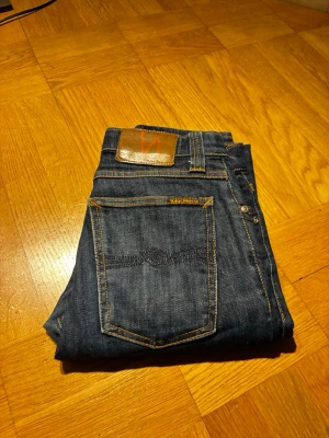 Mörkblå jeans från Nudie Jeans - Säljer ett par mörkblå jeans från Nudie Jeans med klassisk femficksmodell och raka ben. Jeansen har kontrastsömmar och en brun läderpatch med logga bak i midjan. Tillverkade i slitstark denim, perfekta för en clean och stilren look.