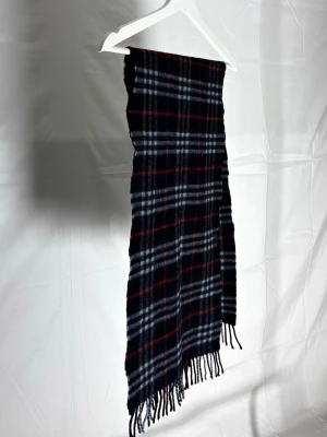 Rutig halsduk i ull från Burberry - Sick 7/10                                                                     Bra sick, inga skador men synlig användning.Mysig halsduk från Burberry i klassiskt rutigt mönster med svarta, röda och vita toner. Halsduken har fransar i ändarna och är tillverkad i mjuk ull som håller dig varm. Perfekt accessoar för att lyfta din outfit med en tidlös look.