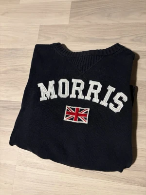 Svart stickad tröja från Morris - Säljer en svart stickad tröja från Morris med vit broderad logotyp och brittisk flagga på bröstet. Tröjan har ribbstickade muddar vid ärmslut och nederkant samt v-ringad hals. Perfekt för dig som gillar klassisk och stilren design.