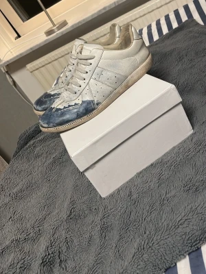 Vita och blå sneakers med läderdetaljer - Säljer ett par vita sneakers med blå detaljer på tån och sidorna. Skorna har klassisk låg profil, perforerade partier för ventilation och vita skosnören. Ovandelen är i läder och sulan är i gummi. Perfekta för dig som gillar en unik och avslappnad stil.Tar byten!