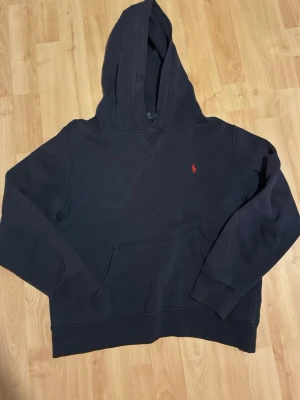 Mörkblå hoodie från Polo Ralph Lauren - Mörkblå hoodie från Polo Ralph Lauren med klassisk huva och känguruficka. Liten röd broderad logga på bröstet. Mjuk bomullskänsla och ribbade muddar vid ärmslut och nederkant. Perfekt för en chill och stilren look.