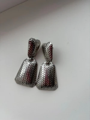 Silvriga geometriska örhängen - Säljer ett par coola silvriga örhängen med geometrisk form och mönstrad yta. Örhängena har en unik, nästan retro vibe och är tillverkade i metall med en snygg glansig finish. Perfekta för att lyfta vilken outfit som helst.
