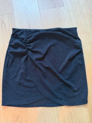 Svart glittrig omlottkjol - Snygg svart kortkjol med glittriga detaljer och omlott-design. Helt oanvänd med tag kvar.