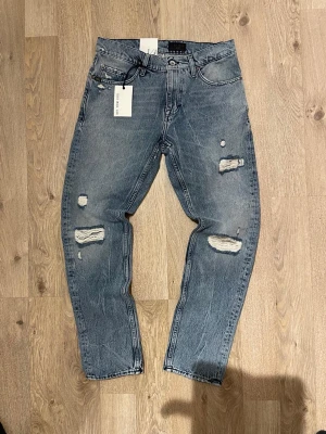 Blå slitna jeans från tiger of sweden - Snygga blå jeans från Tiger of sweden med slitna detaljer och hål på benen. Jeansen har fem fickor, raka ben och klassisk knappgylf. Perfekta för en trendig och avslappnad stil.