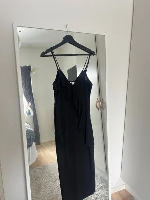 Svart långklänning med volang - Stilren svart långklänning med smala axelband och v-ringning. Klänningen har en snygg volangdetalj framtill som ger en trendig look. Perfekt för dig som gillar minimalistisk stil med en twist. Kanske använd 5 gånger💗