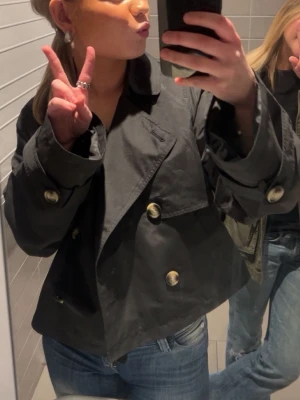 Kort svart trenchcoat med guldknappar - Säljer en trendig kort trenchcoat i svart med stora guldknappar och breda slag. Jackan har en boxig passform och är perfekt att styla med jeans. Materialet ser ut att vara bomull eller polyester.