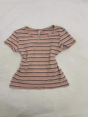 Randig rosa t-shirt från H&M - Snygg rosa t-shirt från H&M med svarta och vita horisontella ränder. Modellen är kortärmad med rund halsringning och har en figurnära passform. Perfekt för en avslappnad och trendig look.