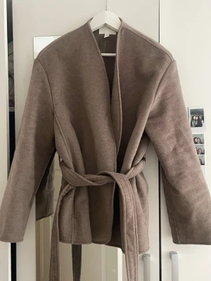 Brun kappa med knytskärp - Stilren brun kappa i mjukt material med knytskärp i midjan och raka ärmar. Kappan har öppen front utan knappar och en minimalistisk design som passar perfekt till lager-på-lager. Perfekt för dig som gillar en clean och modern look.
