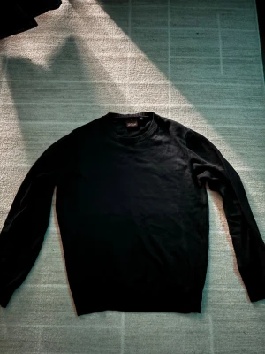 Svart stickad Oscar Jacobson tröja - En stilren svart stickad tröja med rund halsringning och långa ärmar. Tröjan har ribbade muddar vid ärmslut och nederkant, vilket ger en klassisk och enkel look. Perfekt att matcha med jeans för en clean outfit. PRISET KAN DISKUTERAS