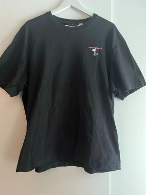 Svart Snoopy t-shirt Beach Beagle - Svart t-shirt med färgglad Snoopy-tryck på ryggen och liten detalj framtill. Rund hals och avslappnad passform. Perfekt för dig som gillar coola och lekfulla motiv.