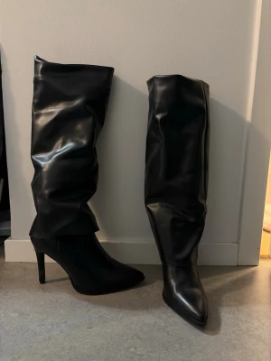 Svarta höga stövlar med klack - Säljer ett par svarta höga stövlar i glansigt konstläder med spetsig tå och smal klack. Stövlarna har en slouchy passform som ger en cool och trendig look. Perfekta för att lyfta vilken outfit som helst.