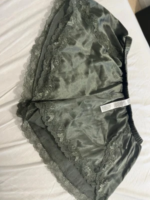 Grå satinskjorts med spets från Censored - Snygga grå satinskjorts från Censored med elastisk midja och dekorativa spetsdetaljer längs kanterna. Perfekta för chill hemma eller som sovplagg. Materialet är mjukt och glansigt för extra komfort.