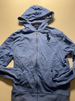 Blå hoodie från Polo Ralph Lauren - Snygg ljusblå hoodie från Polo Ralph Lauren med dragkedja och stor svart logga på bröstet. Hoodien har huva, två fickor framtill och är tillverkad i mjukt bomullsmaterial. Perfekt för en avslappnad och stilren look.