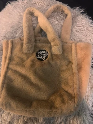 Beige fluffig toteväska med tryck - Supermjuk och fluffig beige handväska i teddy-material med två handtag. På framsidan sitter ett svart och vitt tygmärke med texten 'Today is your day!'. Perfekt för dig som gillar mysiga accessoarer och vill sticka ut lite extra.