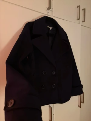 Mörkblå kappa med dubbelknäppning - Stilren mörkblå skepparkavaj med klassisk dubbelknäppning framtill och bred krage. Kavajen har stora knappar både på framsidan och vid ärmsluten. Från design by si, perfekt för kyligare dagar. Enkel men tidlös design som passar till många outfits. Storleken är XS/S 