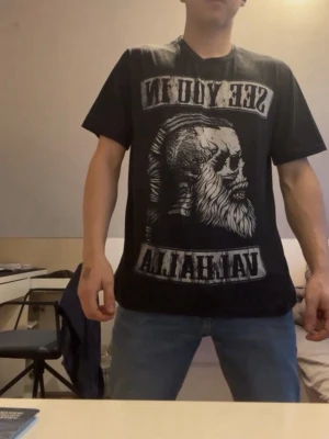 Svart Valhalla t-shirt med tryck - Cool svart t-shirt med stort Valhalla-tryck och motiv av en vikingaskalle både fram och bak. Texten 'SEE YOU IN VALHALLA' ger en edgy vibe. T-shirten har normal passform och är kortärmad, perfekt för dig som gillar streetwear och statement-plagg.