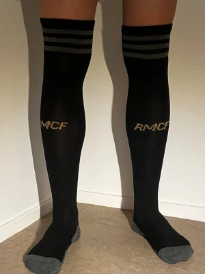 Svarta knähöga strumpor RMCF - Säljer ett par svarta knähöga strumpor från RMCF med grå detaljer på tår och häl. Strumporna har två gråa ränder upptill och gul text på framsidan. Perfekta för dig som gillar sportiga och stilrena accessoarer.