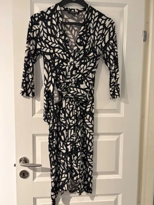 Svartvit omlottklänning med mönster - Snygg svartvit omlottklänning med grafiskt bladliknande mönster. Klänningen har trekvartsärm, v-ringning och knyt i midjan för en smickrande siluett. Perfekt för dig som gillar statementplagg med unik design.