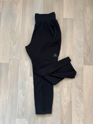 Träningsbyxor från Adidas med dragkedja - Svarta träningsbyxor från Adidas med dragkedjor vid bensluten och fickorna för extra prestanda. Byxorna har en bred resår i midjan och atletisk passform. Diskret Adidas-logga på vänster lår. Byxorna är tillverkade i ett mjukt och stretchigt material vilket gör dem perfekta för träning (t.ex fotboll, gym) eller bara att ha hemma. Använda bara fåtal gånger. Hör gärna av dig vid frågor.