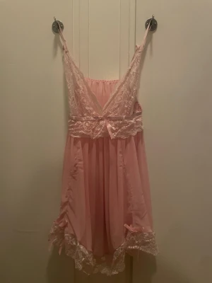 Rosa nattlinne med spets och rosetter - Sött rosa nattlinne med tunna axelband, spetsdetaljer vid byst och nederkant samt små rosetter framtill och vid fållen. Tillverkat i ett mjukt, tunt material som känns luftigt och bekvämt. Perfekt för mysiga kvällar hemma.