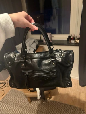 Svart City handväska 🖤 - Säljer en svart handväska i mjukt läder med dubbla handtag och silverfärgade detaljer. Väskan har en dragkedjeficka framtill och klassiska nitar. Perfekt storlek för det viktigaste och ger en cool vibe till din outfit.