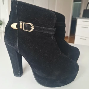 Vintage svarta platåstövletter med gulddetalj - Snygga svarta stövletter i sammet med hög klack och platåsula. 10 cm klack. Dekorativ guldfärgad spänne på sidan och dragkedja bak. Röd sula ger extra edge.   Ca 30 år gamla. Mockan är sliten på vissa ställen inte mer än att den blivit lite blank,  har inte testat att borsta upp. 