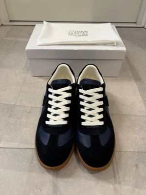 Marin blå sneakers från Maison Margiela - Helt nya snygga marinblå  sneakers från Maison Margiela med vita skosnören och klassisk gummisula. Ovandelen är i en mix av mocka och läder, vilket ger en stilren och exklusiv känsla. 