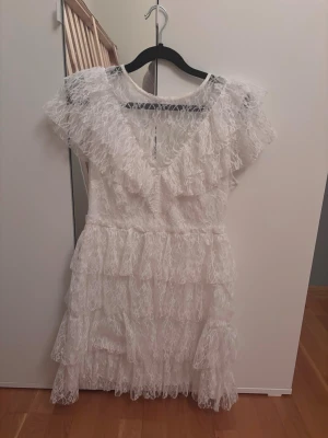 Studentklänning med volanger och spets - Vit klänning med flera lager volanger och spetsdetaljer. Klänningen har korta ärmar och v-ringad halsringning. Perfekt för somriga tillfällen och studentfirande. Använd endast 1 gång (inte på utspring, flak eller utgång, endast på mottagningen). Påminner mycket om klänningarna från ByMalina, men mycket mer prisvärd. 