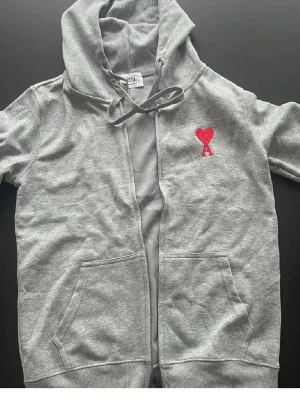 Grå hoodie Ami Paris - Snygg grå hoodie från Ami Paris med dragkedja och kängurufickor. På bröstet finns ett broderat rött hjärta med bokstaven A. Tillverkad i mjuk bomullsmix och har justerbar huva med snören. Perfekt för en chill och stilren look.