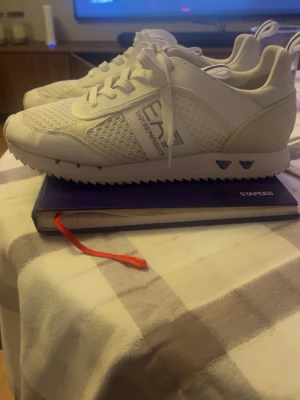 Vita sneakers från Emporio Armani - Snygga vita sneakers från Emporio Armani med meshpaneler och detaljer i syntetläder. Skorna har lågt skaft, vita snören och logga på sidan. Perfekta för en clean och sportig stil.