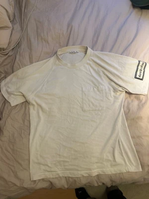 Vit t-shirt från Acne Studios - Vit t-shirt från Acne Studios med bröstficka och patch med logotyp på ena ärmen. Klassisk rund halsringning och rak passform. Materialet är en mix av bomull och polyester, vilket gör den både mjuk och lätt. Perfekt för dig som gillar stilrena basics.