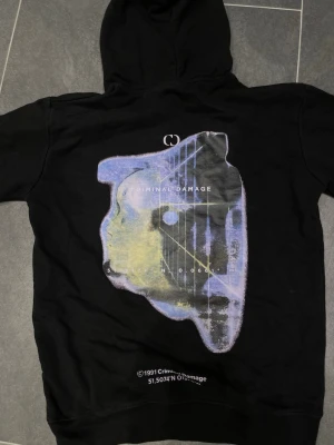 Svart hoodie från Criminal Damage - Svart hoodie från Criminal Damage med stor grafisk print i lila, blått och gult på ryggen samt en mindre färgglad patch på bröstet. Klassisk känguruficka framtill och justerbar huva med snören. Perfekt för dig som gillar streetwear och unika detaljer. Liten fläck på bild 3, syns inte när man har på sig.