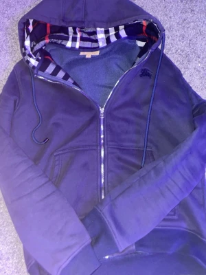 Marinblå hoodie från Burberry - Snygg marinblå hoodie från Burberry med dragkedja och klassiskt rutigt foder i hu for extra stilpoäng. Huva med dragsko och två fickor framtill. Perfekt för dig som vill ha en exklusiv och bekväm look. Materialet känns mjukt och kvalitativt.