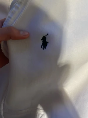 Vit hoodie polo Ralph Lauren - Säljer en klassisk vit hoodie från Polo Ralph Lauren med dragkedja och den ikoniska broderade loggan på bröstet. Hoodien har en mjuk bomullskänsla och ribbade muddar. Perfekt för en clean och stilren look.