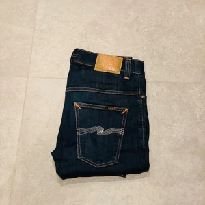 Nudie jeans - Säljer ett par mörkblå jeans från Nudie Jeans med klassisk femficksmodell och kontrastsömmar. Jeansen har en snygg bakficka med vågig söm och patch i läderimitation vid linningen. Tillverkade i slitstark bomull med en modern passform.
