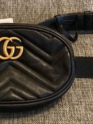 Svart magväska  - Snygg svart magväska m logga i guld framtill och hjärtdetalj på baksidan. Väskan har justerbart bälte och oval form, perfekt för att bära det viktigaste med stil. I mycket fint skick!