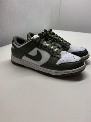 Nike Dunk Low olivgrön/vit - Nike Dunk Low sneakers i olivgrönt och vitt läder med klassisk Swoosh på sidan. Skorna har perforerad tåbox, vita paneler och olivgröna detaljer samt matchande snören. Yttersulan är i gummi och designen är tidlös och clean.
