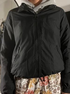 Svart vindjacka med dragkedja - Stilren svart vindjacka med hel dragkedja framtill och två snedställda fickor med dragkedja. Jackan har en enkel design och en luva som skyddar mot vind. Perfekt för blåsiga dagar och enkel att matcha med olika outfits.