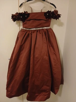 Brun barn långklänning med blommor - Snygg brun barn långklänning med offshoulder sopar och stora dekorativa blommor på axlarna. Klänningen har en glittrig bård i midjan och är tillverkad i ett glansigt tyg som ger en elegant look. Perfekt för speciella tillfällen.