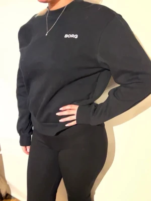 Svart sweatshirt från BORG - En stilren svart sweatshirt från BORG med rund halsringning och diskret logga på bröstet. Tröjan har långa ärmar och ribbade muddar vid ärmslut och nederkant. Perfekt till ett par tights eller jeans för en clean look.