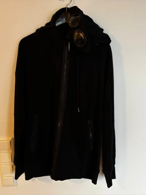 Svart hoodie med glasögon från C.P. Company - Säljer en svart hoodie från C.P. Company med ikoniska inbyggda glasögon i huvan. Hoodien har dragkedja framtill, två fickor med dragkedja och ribbade muddar. Tillverkad i mjukt bomullsmaterial och har en modern, avslappnad passform. Adiatiskt XXL, som europesk L. Ny.