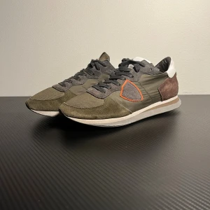 Gröna sneakers från Philippe Model - Snygga sneakers från Philippe Model i grönt och grått med orange detalj på sidan. Skorna har en mix av mocka och textil, vit häl och bruna partier bak. Klassisk snörning och platt sula, perfekt för dig som gillar streetstyle med en unik touch.