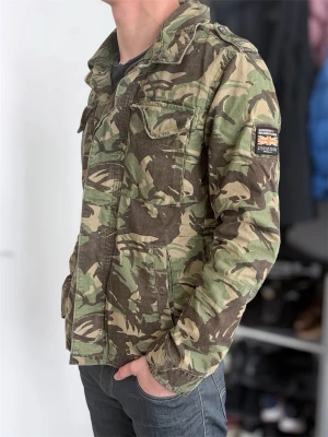 Camouflage overshirt från Superdry - Säljer en grön och brun camouflage overshirt från Superdry med klassisk krage, bröstfickor och patch med brittisk flagga på ärmen. Jackan har knappar framtill och är perfekt för dig som gillar streetwear och utility-vibe.