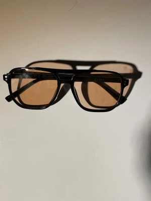 Svarta solglasögon med bruna glas - Snygga solglasögon med svart, kantig båge och bruna tonade glas. Klassisk pilotform som ger en cool retrovibe. Perfekta för soliga dagar och en stilren look.