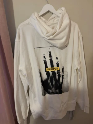 Vit hoodie från Purple brand  - Säljer en vit hoodie från Purple med stor ficka framtill och huva med dragsko. På bröstet står det 'PURPLE' i svart text och på ryggen finns ett coolt tryck av en röntgenhand med gul text. Aldrig använd. Nypris 2800kr