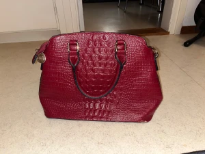 Vinröd handväska med krokopräglat mönster - Snygg vinröd handväska i konstläder med krokopräglat mönster och guldfärgade detaljer. Väskan har två handtag och dragkedja upptill samt dekorativt hänge i metall. Perfekt för dig som vill ha en trendig accessoar med lyxig känsla.En liten fläck (se på sista bilden)
